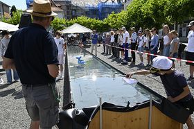 Menschen stehen an einem langen, schmalen Wasserbecken im Freien und beobachten ein kleines blaues, walförmiges Modellboot im Wasser. Im Vordergrund ein Mann mit Strohhut, rechts ein Junge mit Cap, der sich über das Becken beugt.