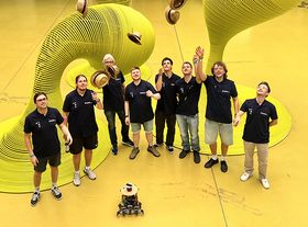 Acht Personen mit dunkelblauen Shirts werfen einen Strohhut in die Höhe. Vor ihnen steht ein mobiler Roboter. Im Hintergrund sind große gelbe astrakte Figuren auf einem gelben Boden platziert.