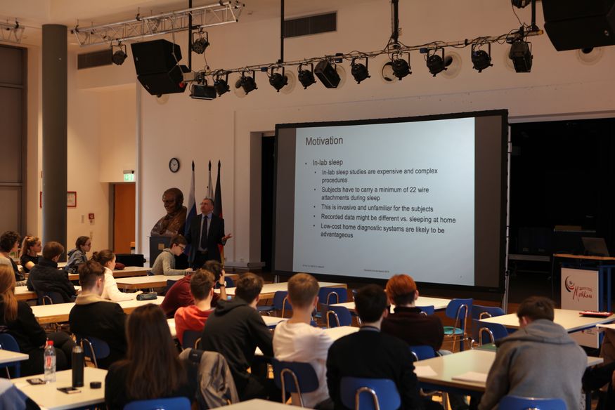 Prof. Dr. Ralf Seepold steht in einer Klasse vor Schülern neben einer weißen Leinwand, auf der schwarze Schrift zu erkennen ist.