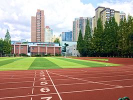 Der Sportplaz auf dem Campus der Shanghai Jiaotong Universität an einem sonnigen Tag. Man sieht grünen Rasen vor Gebäuden in der Ferne und eine rotbraune Laufbahn mit Zahlenmarkierungen, die um die Grünfläche herumführt. 