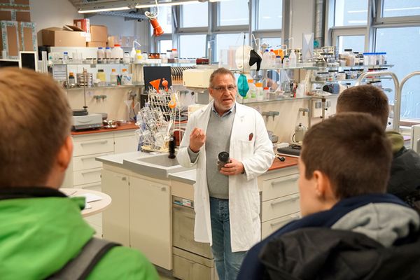 Laboringenieur Manfred Schollenberger steht in einem weißen Kittel zwischen Laborschränken und blickt in die Kamera. Von hinten sind die Rücken von Schülern zu sehen, die im Halbkreis um ihn stehen.