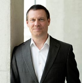Ein Mann trägt weißes Hemd und schwarzes Jacket und lächelt freundlich in die Kamera