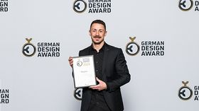 Aric Merz trägt schwarzen Anzug und steht vor einer Fotowand. In der Hand hält er die Urkunde über den German Design Award