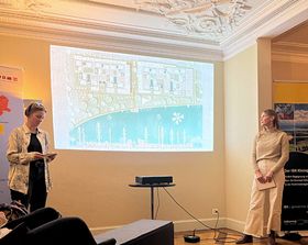 Das Bild zeigt zwei Studentinnen, die eine Präsentation halten. An die Wand hinter ihnen ist das Bild eines Architekturentwurfs projeziert.