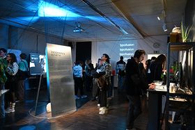 Ein blau ausgeleuchteter Ausstellungsraum mit Screens und Texttafeln, in dem sich ein gemischtes Publikum die Exponate ansieht