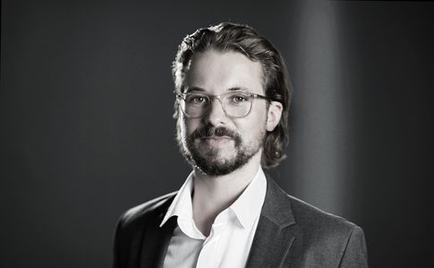 Schwarz-Weiß-Bild von Dr. Marcus Ehrenberger. Er hat die mittellange Haare zurück gekämmt, trägt einen Bart und Brille zu dem dunklen Anzug.