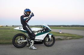 Ein Motorradfahrer im Rennanzug mit Helm posiert auf einem Motorrad auf einer Rennstrecke fürs Foto. Er reckt den Daumen in die Luft.
