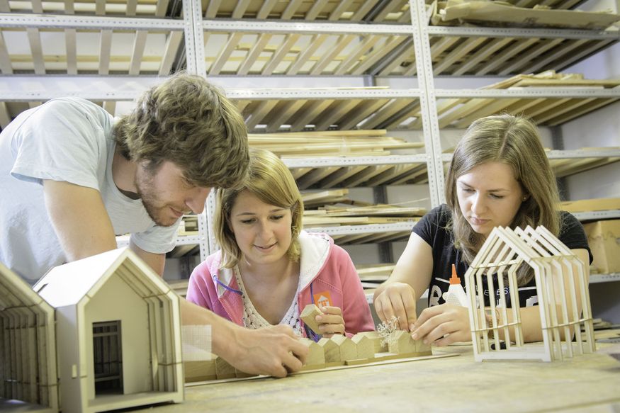 Zwei Frauen sitzen an einem Tisch und betrachten Architekturmodelle, die auf dem Tisch stehen. Ein junger Mann beugt sich über sie und berührt eines der Modelle.