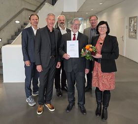 Das Bild zeigt fünf Männer in dunklen Anzügen und eine Frau in rotem Kleid und Blazer. In der Mitte des Gruppe hält ein Mann eine Urkunde stolz in den Händen, die Frau hält einen Blumenstrauß. Alle lächerln in die Kamera.