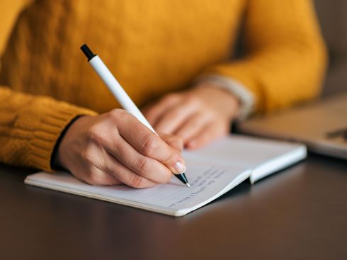 Eine junge Frau im gelben Pullover schreibt in ein Notizbuch. Anonymer Ausschnitt.