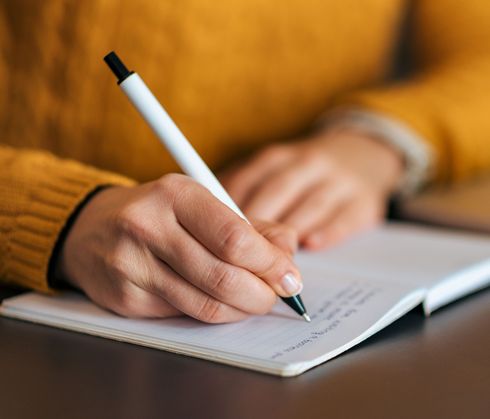 Eine junge Frau im gelben Pullover schreibt in ein Notizbuch. Anonymer Ausschnitt.