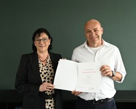 Ein Mann und eine Frau halten eine Urkunde in der Hand und lächeln in die Kamera