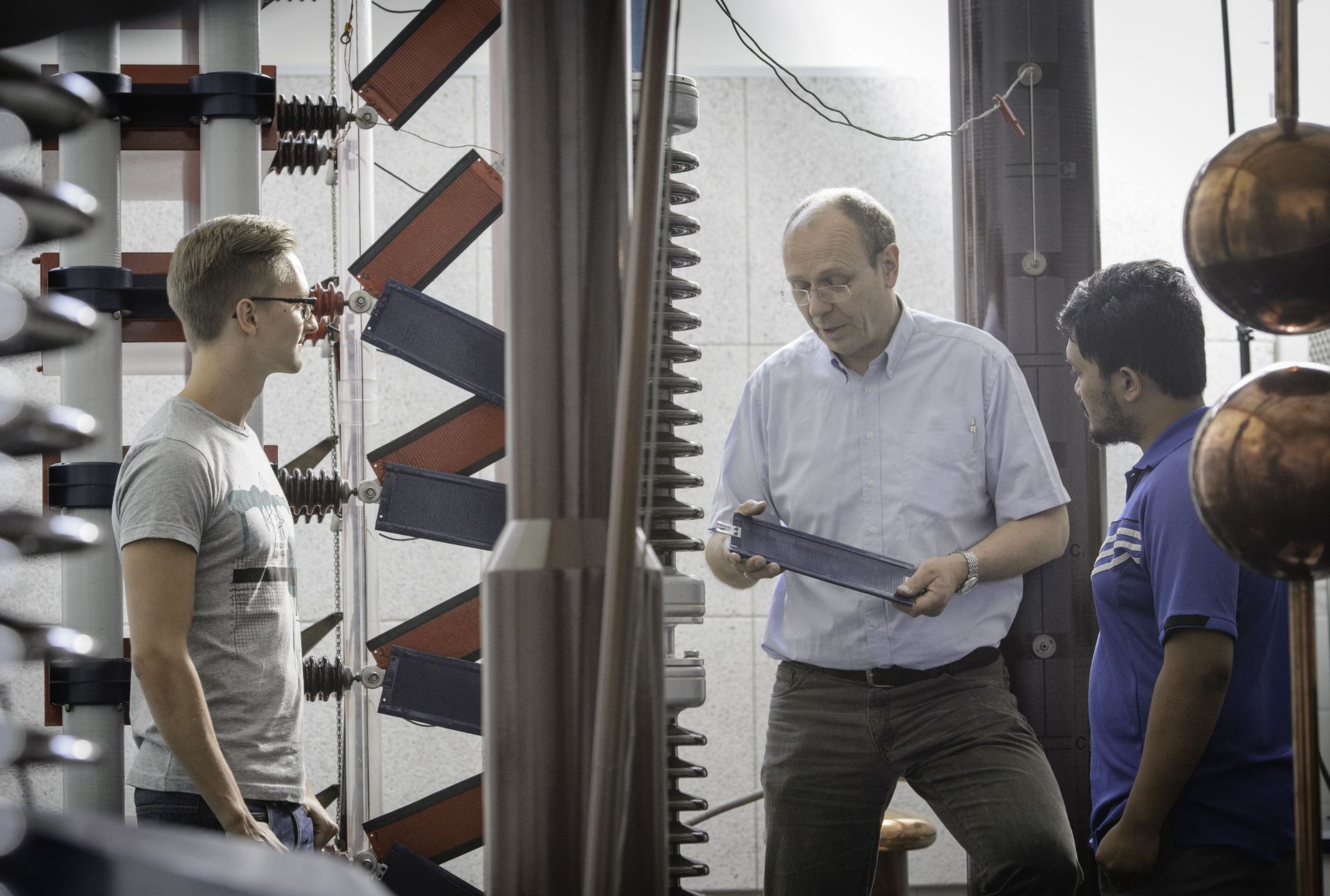 Ein Professor zeigt zwei Studierenden Bauteile einer Maschine