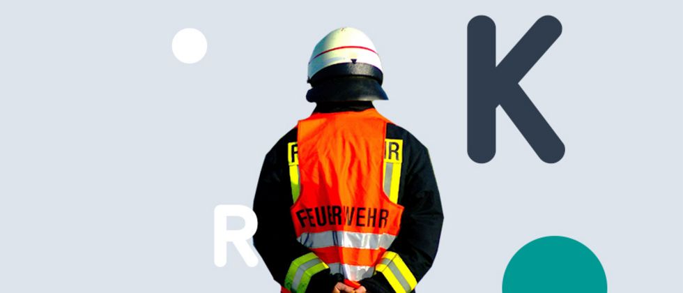 Man sieht den Rücken eines Feuerwehrmannes in roter Warnweste und weißem Helm vor einer grauen Fläche. Darauf befinden sich die die Buchstaben K und R.