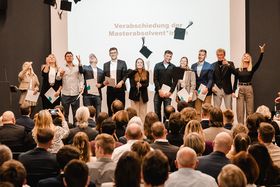 Das Bild zeigt eine Gruppe von Absolventinnen und Absolventen auf einer Bühne. Sie werfen fröhlich ihren Absolventenhut in die Luft.