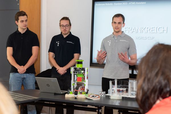 Drei Studenten stehen hinter einem Tisch, auf dem ihre Roboter stehen.