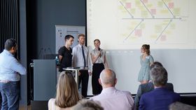Blick in einen Vortragssaal. Zu sehen sind die Rücken von Gästen, die im Auditorium sitzen. Auf der Bühne stehen fünf Studentinnen und Studenten vor einer Präsentationsfolie.