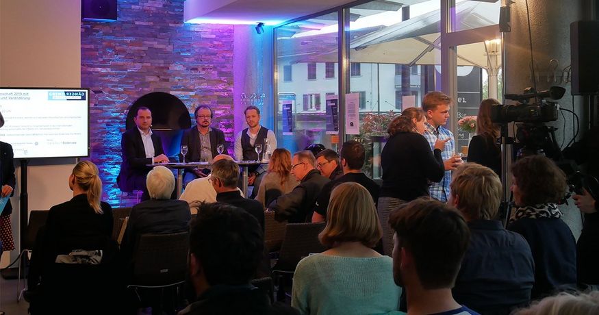 In einem lila und blau illuminiertem Café sitzen vier Männer auf Barhockern und blicken in ein Publikum.