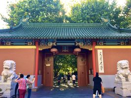 Ein traditionelles chinesisches Gebäude aus rotem Gestein mit bunt bemalter Abschlusskante, über der ein Dach aus dunkelgrünen Dachpfannen thront. Es handelt sich um einen Durchgang zum Park. An der Mauer neben dem Durchgang hängt in chinesischen Zeichen im Kalligrafie-Stil der Name der Universität: Jiaotong Daxue. Hinter dem Durchgang sieht man grüne Baumkronen. Vereinzelt sind chinesische Passanten zu sehen. 