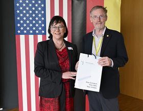 Das Bild zeigt Sabine Rein und Robert McMahan vor der US-amerikanischen und deutschen Flagge stehend. Rein überreicht McMahan eine Geschenktüte 