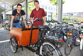 Ein Lastenfahrrad steht vor weiteren abgestellten Fahrrädern. Ein junger Mann lehnt sich auf die Transportkiste und lächelt in die Kamera. Ein weiterer steht daneben und schaut ebenfalls in die Kamera.