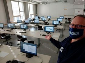 Blick aus der Vogelperspektive in einen Seminarraum mit rund 20 aktivierten Bildschirmen. Recht im Bild ist Laboringenieur Ingo Kletti mit Mund-Nasen-Bedeckung zu sehen. Er zeigt mit seinem rechten Arm in den Raum.