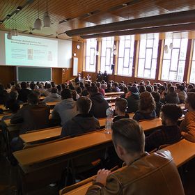 Blick von hinten nach vorn in einen mit Menschen gefüllten Raum, die an Tischreihen sitzen, vorn ist eine Leinwand sowie eine angeschnittene Tafel zu sehen