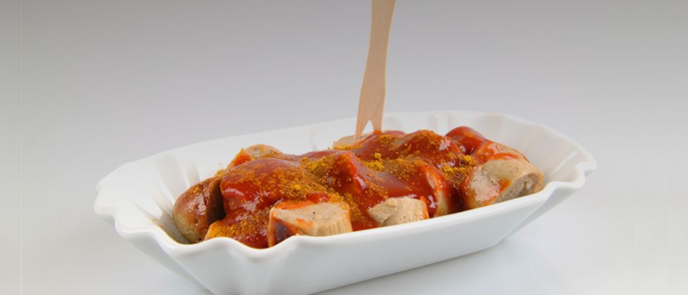 In einem Pappschälchen befindet sich eine Currywurst mit einem Holzspieß.