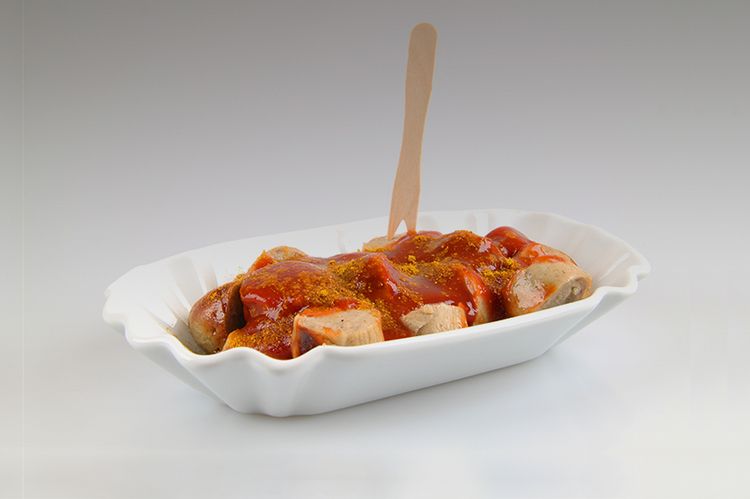 In einem Pappschälchen befindet sich eine Currywurst mit einem Holzspieß.