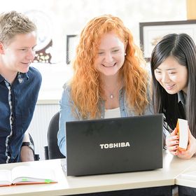 Das Bild zeigt drei Studierende, die gemeinsam auf den Laptop der Studentin in der Mitte schauen. Sie lachen
