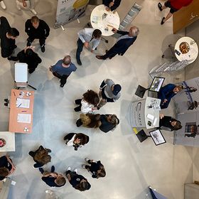Eine Menschenmenge von oben. Einige Personen stehen vor Bannern und diese scheinen im Kontakt mit Personen zu seinen, die vor ihren Tischen stehen.