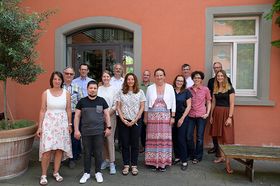 Das Bild zeigt eine dreizehnköpfige Gruppe von Frauen und Männern, die vor einer organgefarbenen Wand im Freien stehen. 