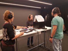 Szene im Tonstudio. Um einen Tisch stehen von links Theaterpädagogin Anna Hertz, Studentin Janina, Schauspieler Thomas Fritz Jung. Sie alle tragen Kopfhörer und blicken auf Textseiten vor sich.
