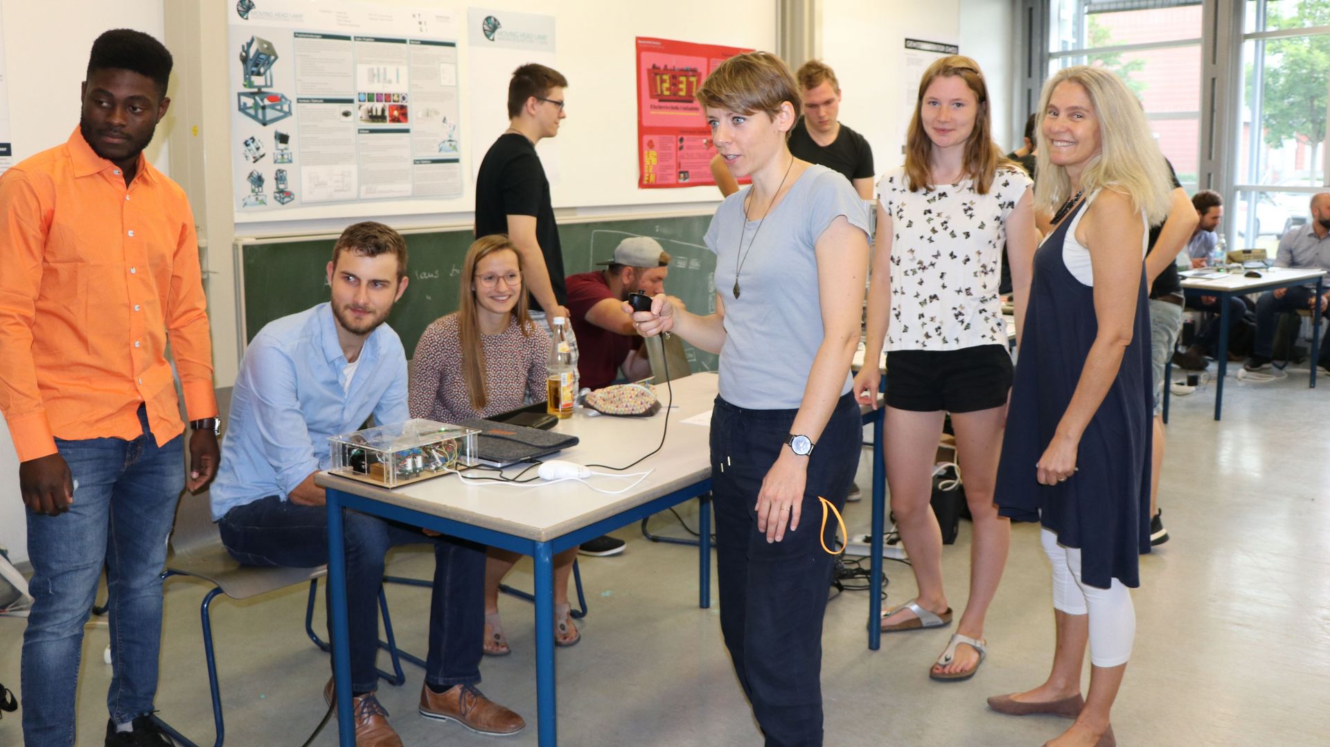 Studierende sitzen an ihrem Ausstellungstisch mit ihrem selbst gebauten Exponat, im Vordergrund testen Fakultätsreferentinnen von EI das Exponat.