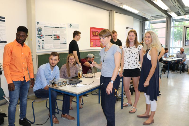 Studierende sitzen an ihrem Ausstellungstisch mit ihrem selbst gebauten Exponat, im Vordergrund testen Fakultätsreferentinnen von EI das Exponat.