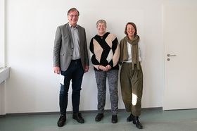 Gruppenfoto vor einer weißen Wand, von links nach rechts: Thomas Birkhölzer trägt eine schwarze Jeans, graues Hemd und Jackett. In der Hand hält er den Kooperationsvertrag. In der Mitte steht Hanna Schönfeld, sie trägt eine graugemusterte Hose und einen schwarz-beige gemusterten Strickpullover, rechts steht Sandra Flügel in khakifarbener Hose und Schal und weißem Shirt.