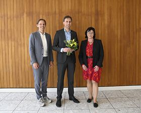 Zwei Männer und eine Frau stehen bei einer Gratulation vor einer Wand