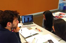 Vier Studierende bei einer Videokonferenz: Sie sitzen an einem Tisch, auf dem ein Laptop steht und schauen auf den Bildschirm. Darauf zu sehen sind drei weitere Personen.