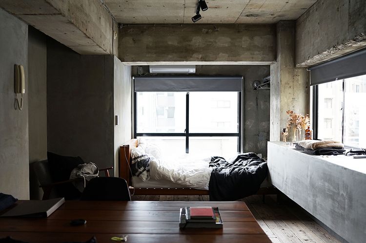 Blick in ein Zimmer: Im Vordergrund steht ein Tisch, vor dem Fenster am gegenüberliegenden Ende des Raumes steht ein Bett.