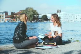 Zwei Studentinnen sitzen am Seehrein, lernen gemeinsam. Im Hintergrund der Campus der HTWG.
