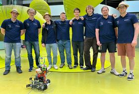Acht Personen mit dunkelblauem Polo und Strohhut stehen nebeneinander, vor ihnen steht ein Roboter.