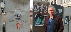 Mann mit grauen Haaren, Ende 50, trägt ein braunes Jackett und steht vor einer Betonwand mit Grafitti.