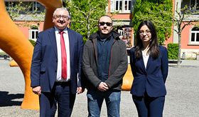 Zwei Männer und eine Frau lächeln in die Kamera, im Hintergrund ein Kiesplatz mit einer organgenen Skulptur und ein mit grün bewachsenes Gebäude