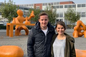 Eine junge Frau und ein junger Mann posieren Schulter an Schulter vor den orangenen Symbolfiguren auf dem HTWG-Campus.