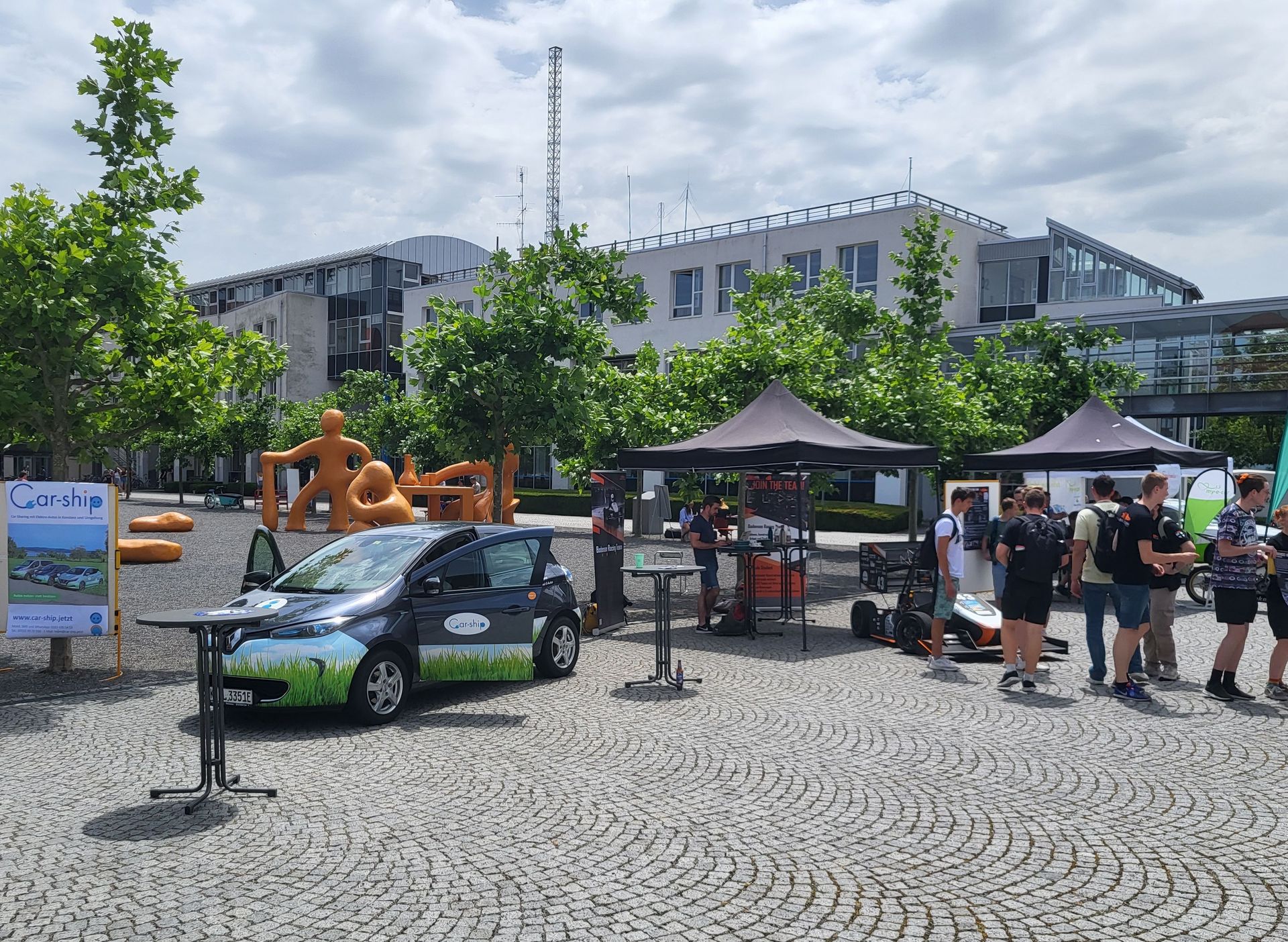 Foto mit Personen an Ständen des Mobilitätsmittwochs. Dabei stehen Menschen um Pavillons und ein Elektroauto