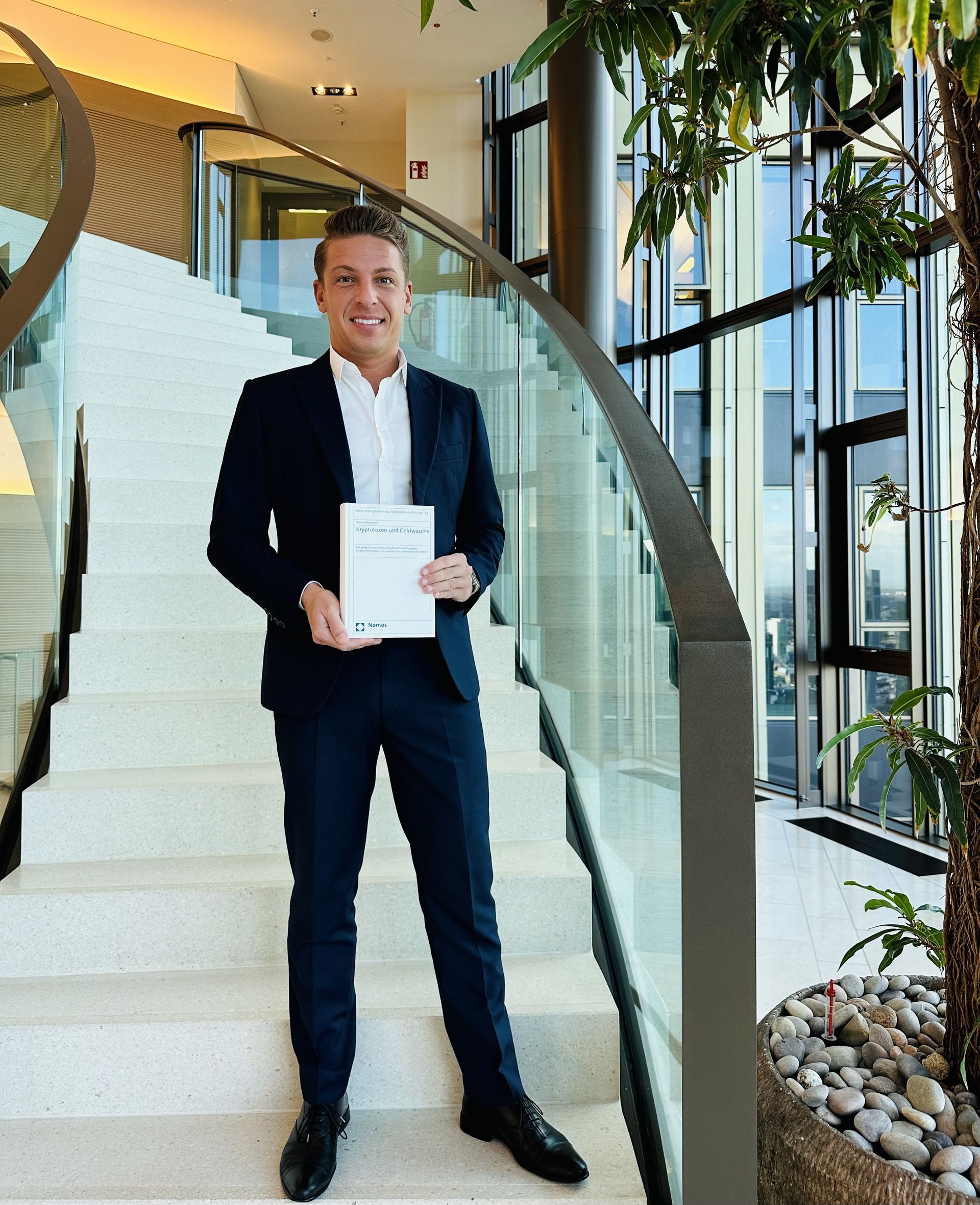 Dr. Simon Biberacher mit einer Druckausgabe seiner Doktorarbeit am Fuße einer Treppe stehend