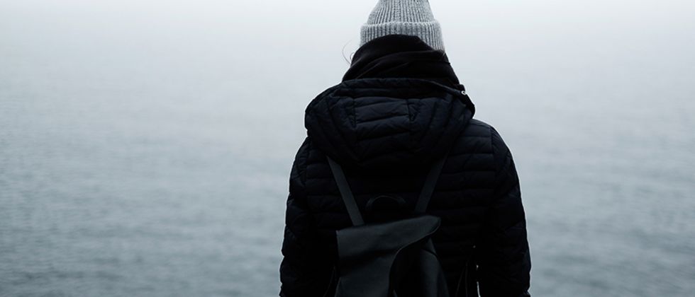 Eine Frau in dicker dunkler Jacke, schwarzem Schal und grauer Mütze mit einem schwarzen Rucksack steht mit dem Rücken zum Betrachter des Bildes vor einem See. Sie scheint den Kopf hängen zu lassen.