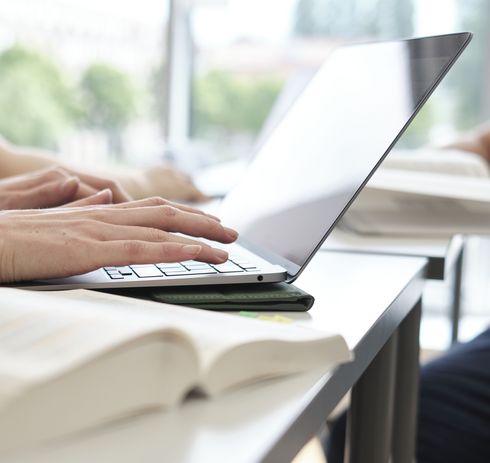 Ausschnitt eines Arbeitsplatzes mit Laptop, Händen auf der Tastatur, aufgeschlagenem Buch.