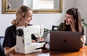 Zwei junge Frauen sitzen an einem Tisch und lächeln sich an. Vor der einen steht eine Nähmaschine, vor der anderen ein Laptop.