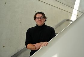 Ein junger Mann mit locken und Brille lehnt an einem Treppenaufgang